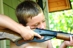 Picture of a little blond boy with a bb gun, aiming it outside from a doorway