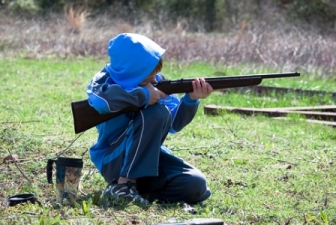 Photo of a boy in blue jacket and hood holding and aiming an airsoft rifle gun outside in the grass fields. Is airsoft dangerous? Not if you teach your children gun safety.